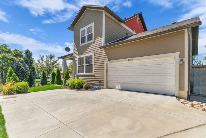 Exterior siding finish, white paneled garage door, concrete driveway, mature landscaping, and an exterior wall light fixture