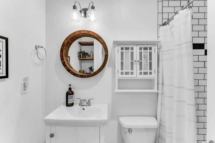 White subway tile shower surround with a black accent band, a white vanity with an integrated sink, a circular wood-framed mirror, and a wall-mounted storage cabinet