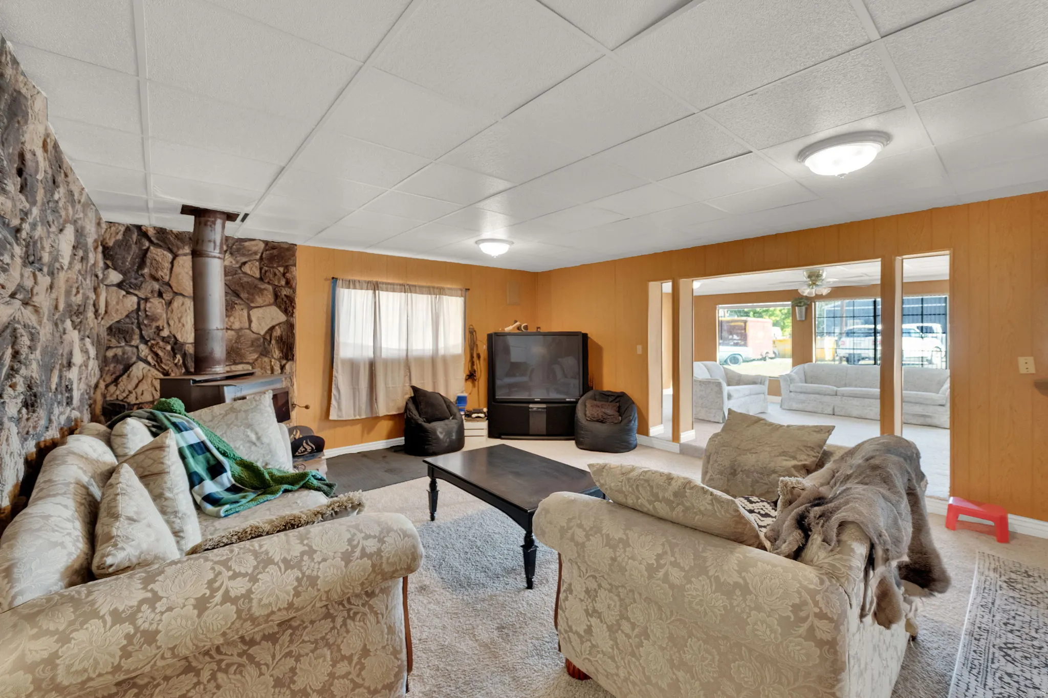 Carpeted living room with a wood stove, a paneled ceiling, wood walls, and a ceiling fan