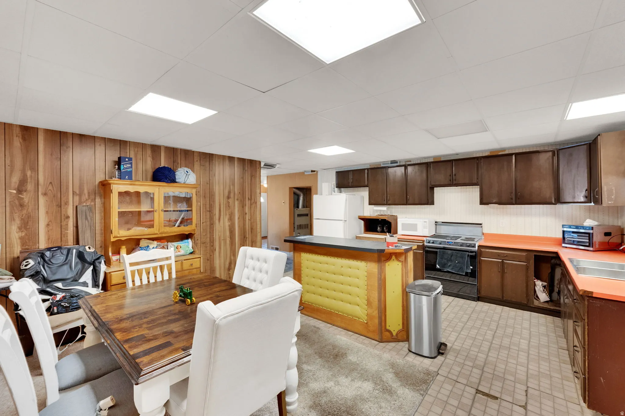 Kitchen featuring dark wood finish cabinetry, white appliances, a kitchen island, wood walls, and a drop ceiling