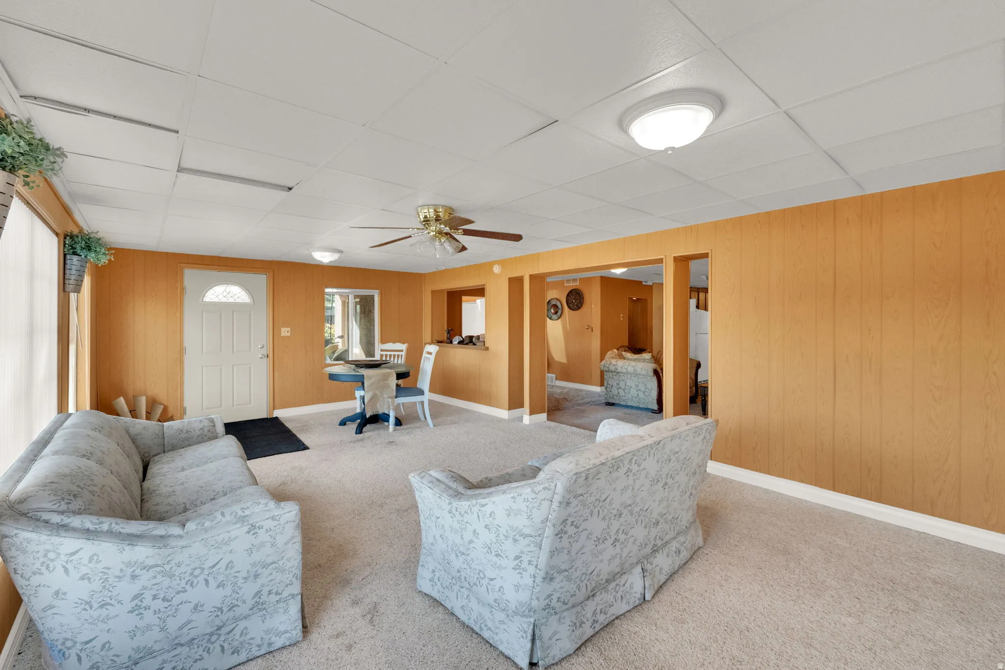 Living area featuring a ceiling fan, wooden walls, carpet, and a drop ceiling