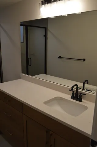 Wood-finish vanity with drawer storage, integrated sink, light-toned countertop with backsplash, matte black faucet, and a wall-mounted mirror with an overhead light fixture