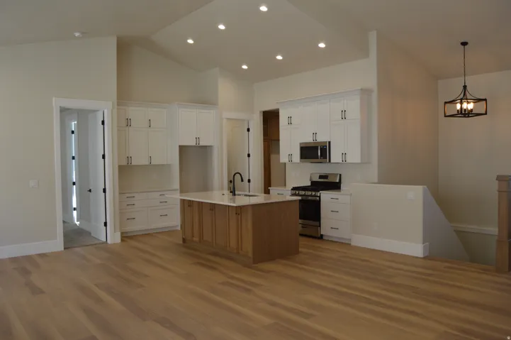 Open-concept kitchen and living area featuring wood-finish flooring, vaulted ceilings, recessed lighting, white cabinetry with black hardware, and a center island with a sink