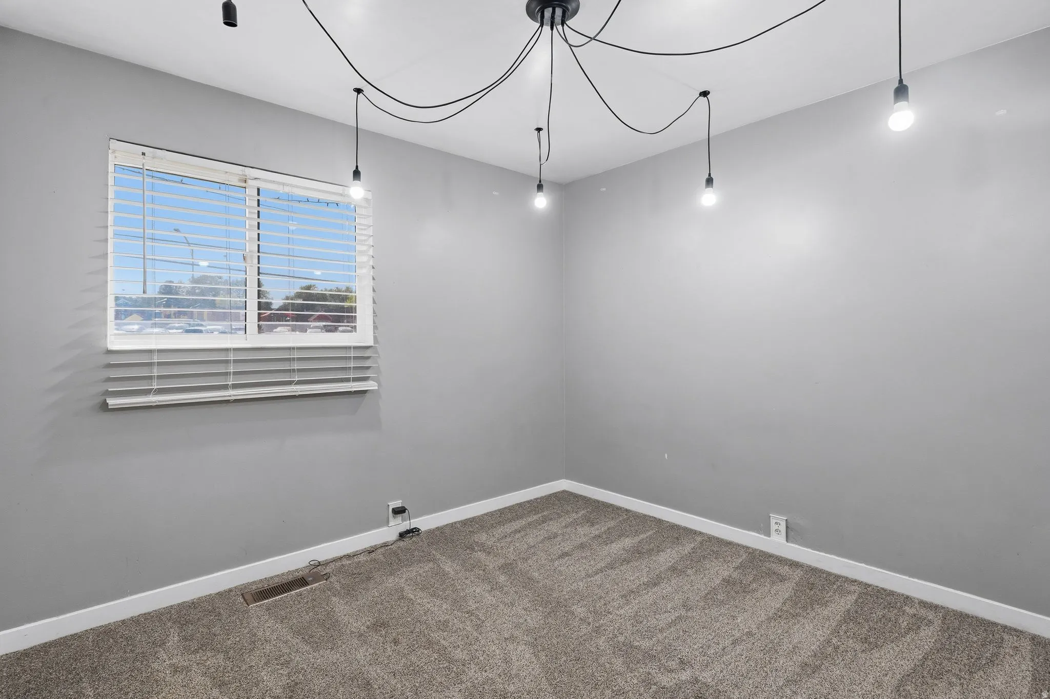 Bedroom featuring a window with white blinds, contemporary ceiling light fixture with exposed bulbs, neutral grey walls, and plush carpet flooring