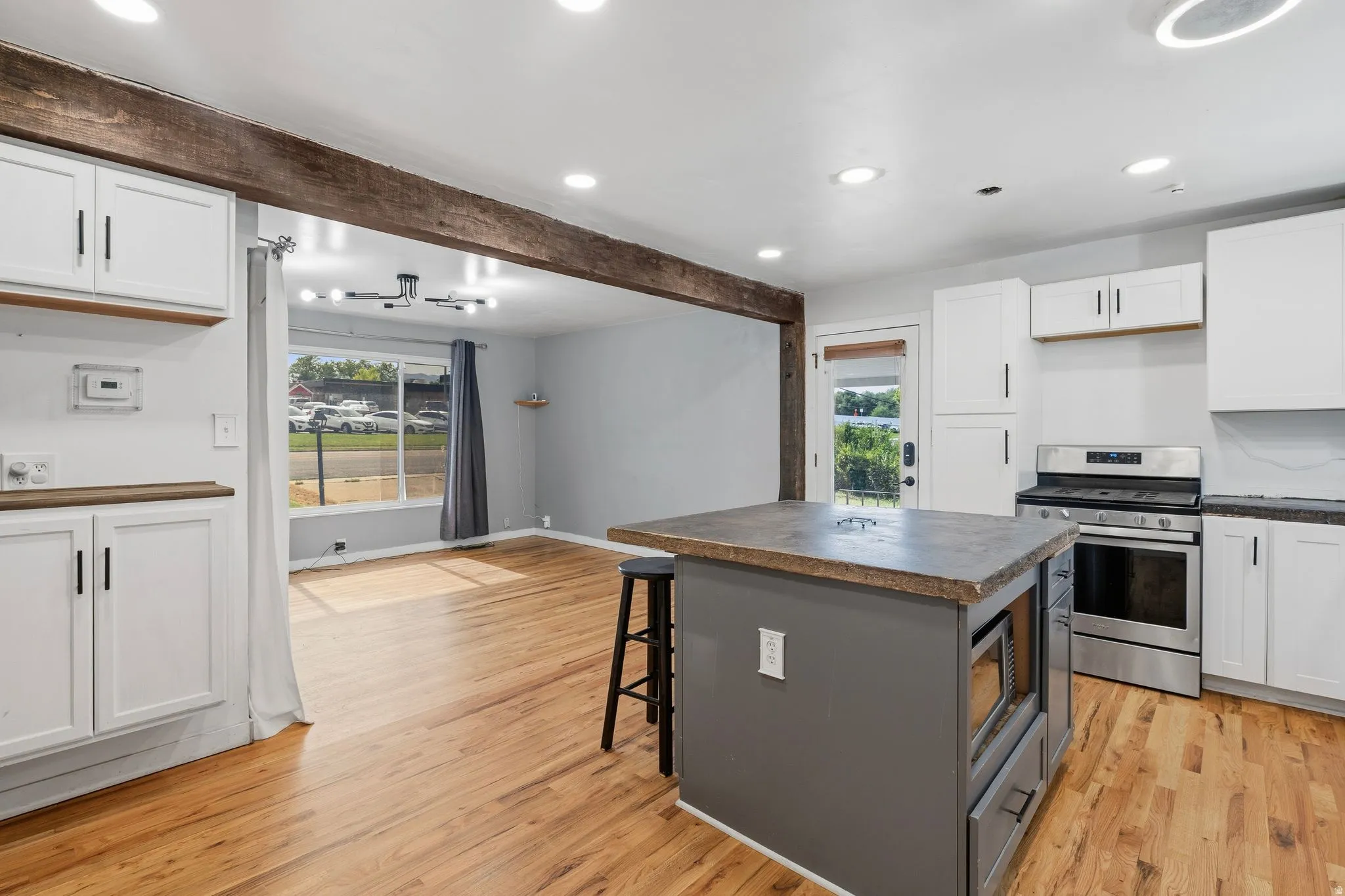Open-concept kitchen and living area featuring a rustic wood beam, light wood-finish flooring, white cabinetry with dark hardware, stainless steel appliances, and a kitchen island with a built-in microwave