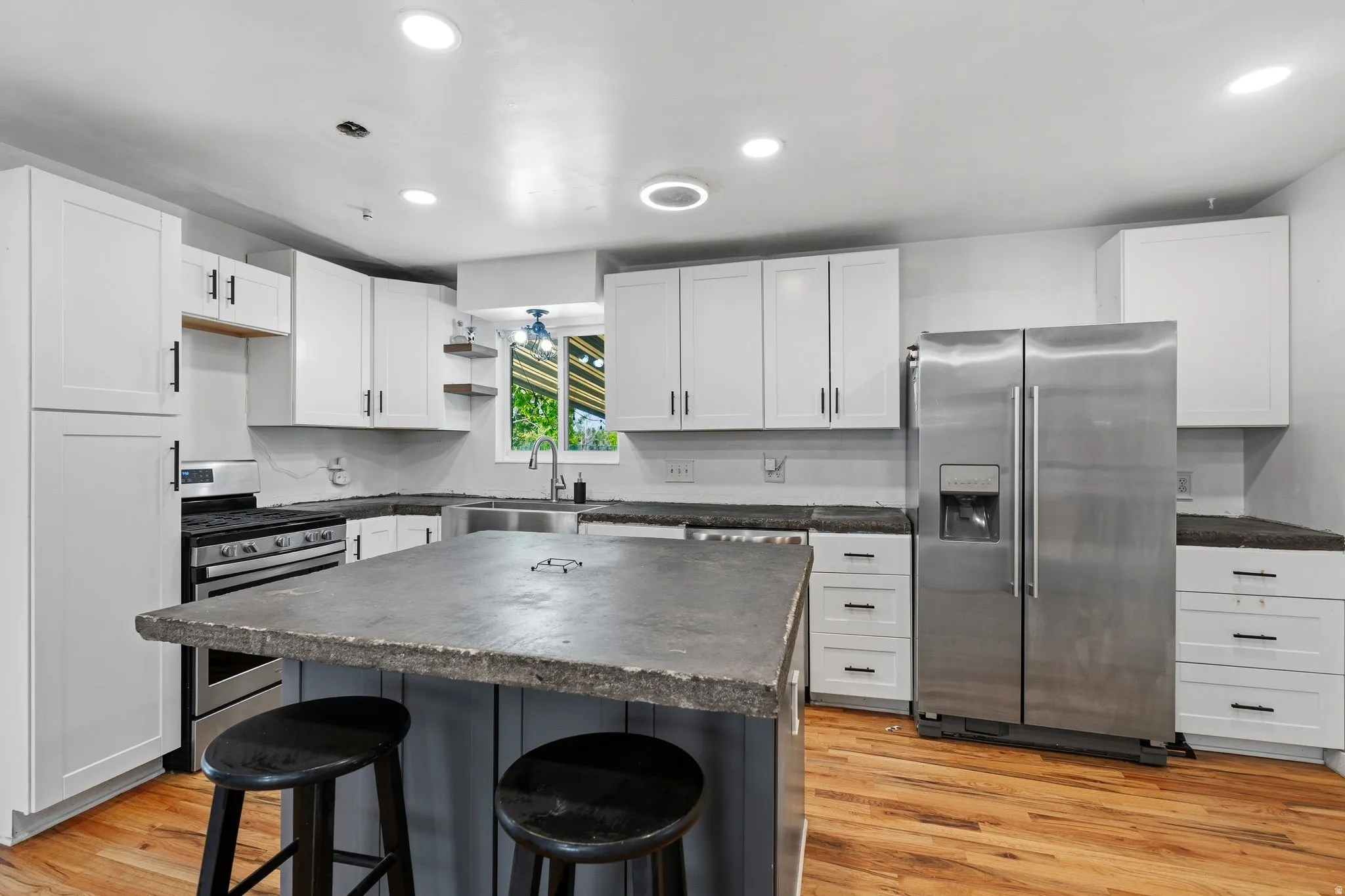 Spacious kitchen featuring an oversized island with a dark stone-look countertop, white shaker cabinetry, stainless steel appliances, recessed lighting, and wood-finish flooring