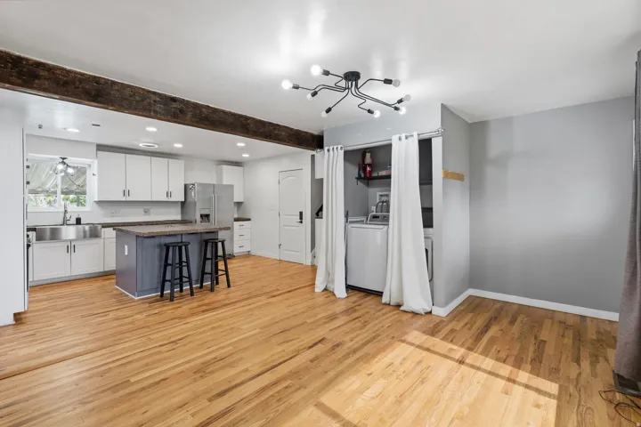 Open-concept living area featuring wood-finish flooring, a contemporary ceiling fixture, a kitchen with white cabinetry and a central island, and a dedicated laundry alcove