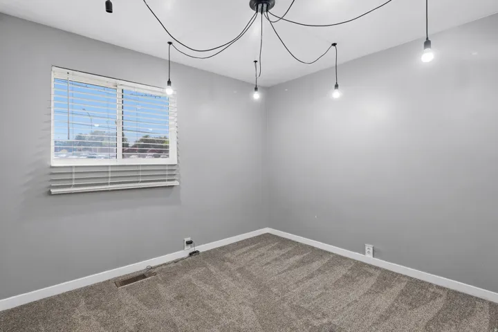 Bedroom featuring a window with white blinds, contemporary ceiling light fixture with exposed bulbs, neutral grey walls, and plush carpet flooring