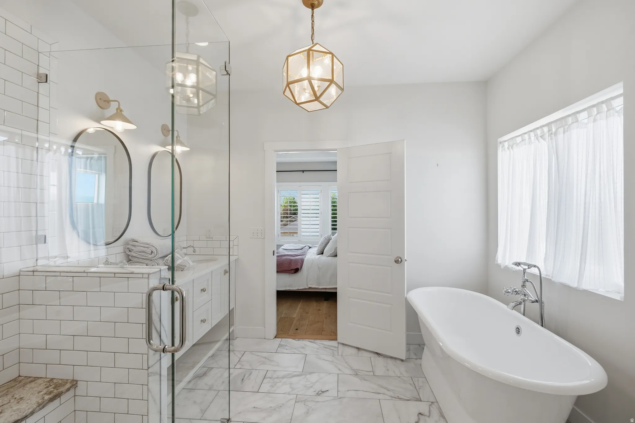 Spacious bathroom featuring a freestanding soaking tub, large walk-in shower with a built-in bench, double vanity with dual oval mirrors, marble-look tile flooring, and a unique geometric pendant light fixture