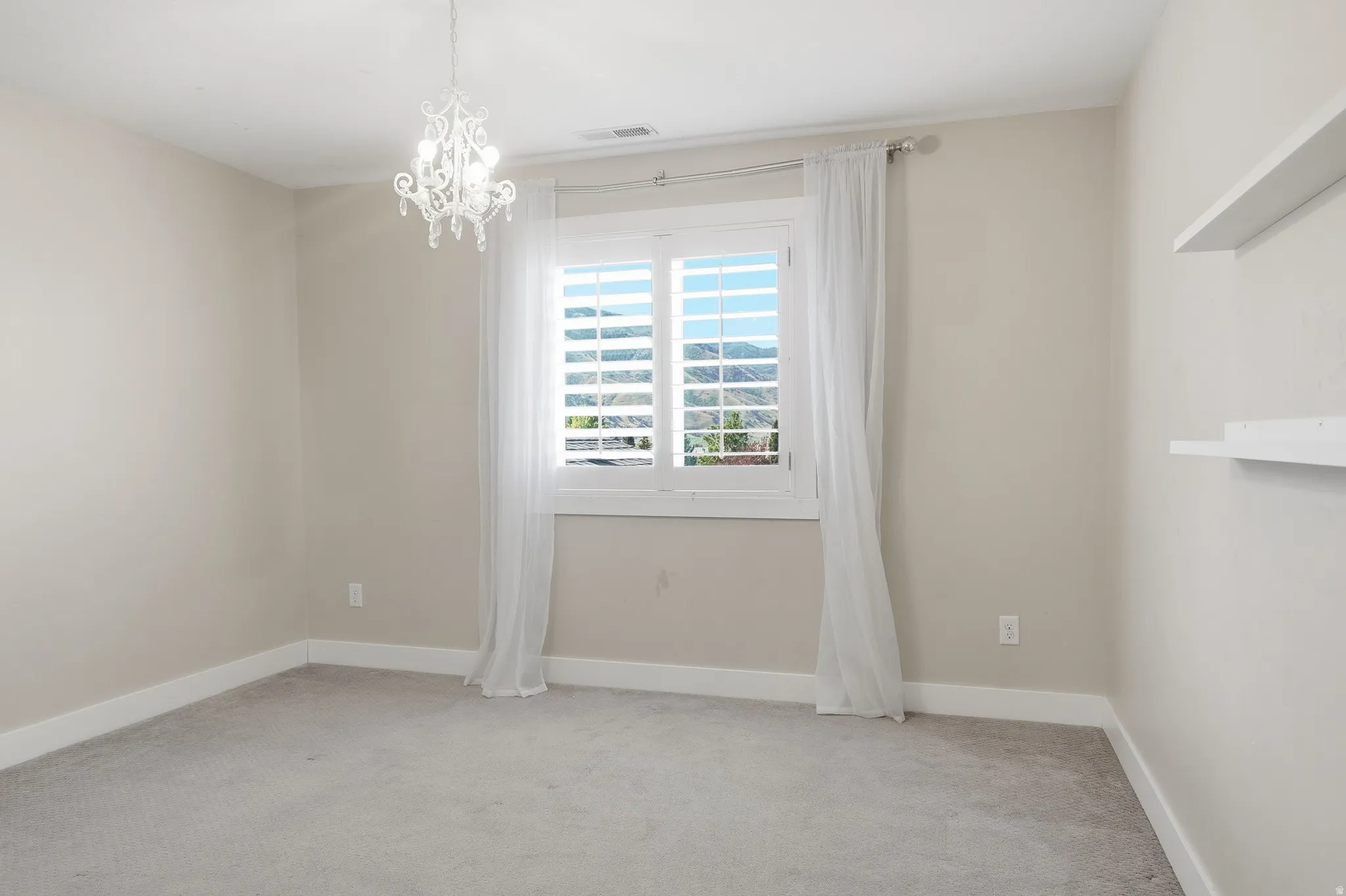 Room featuring a decorative chandelier, plantation shutters, white window sheers, light gray carpeting, and integrated wall shelving