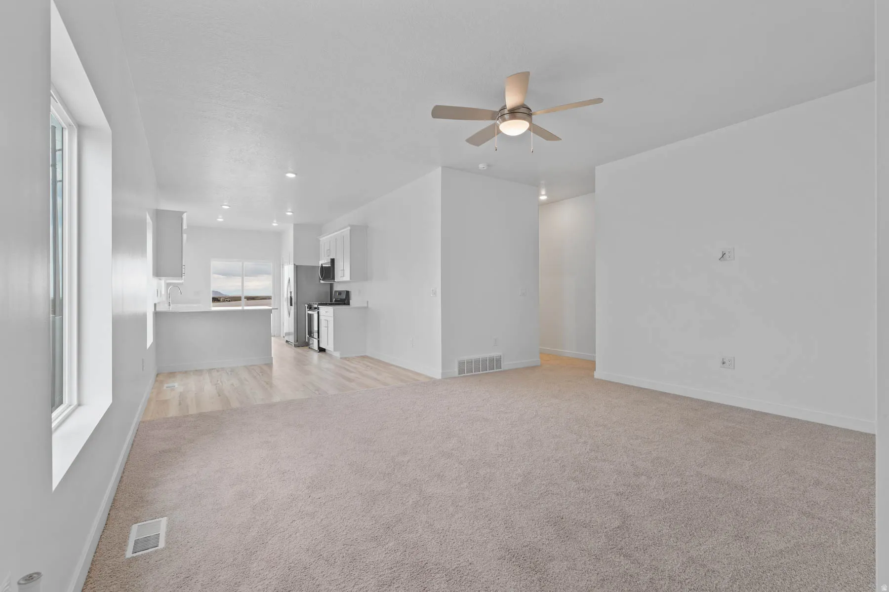 Open-concept living area with neutral carpeting and a ceiling fan, featuring a kitchen with white cabinetry, stainless steel appliances, and a peninsula with a sink