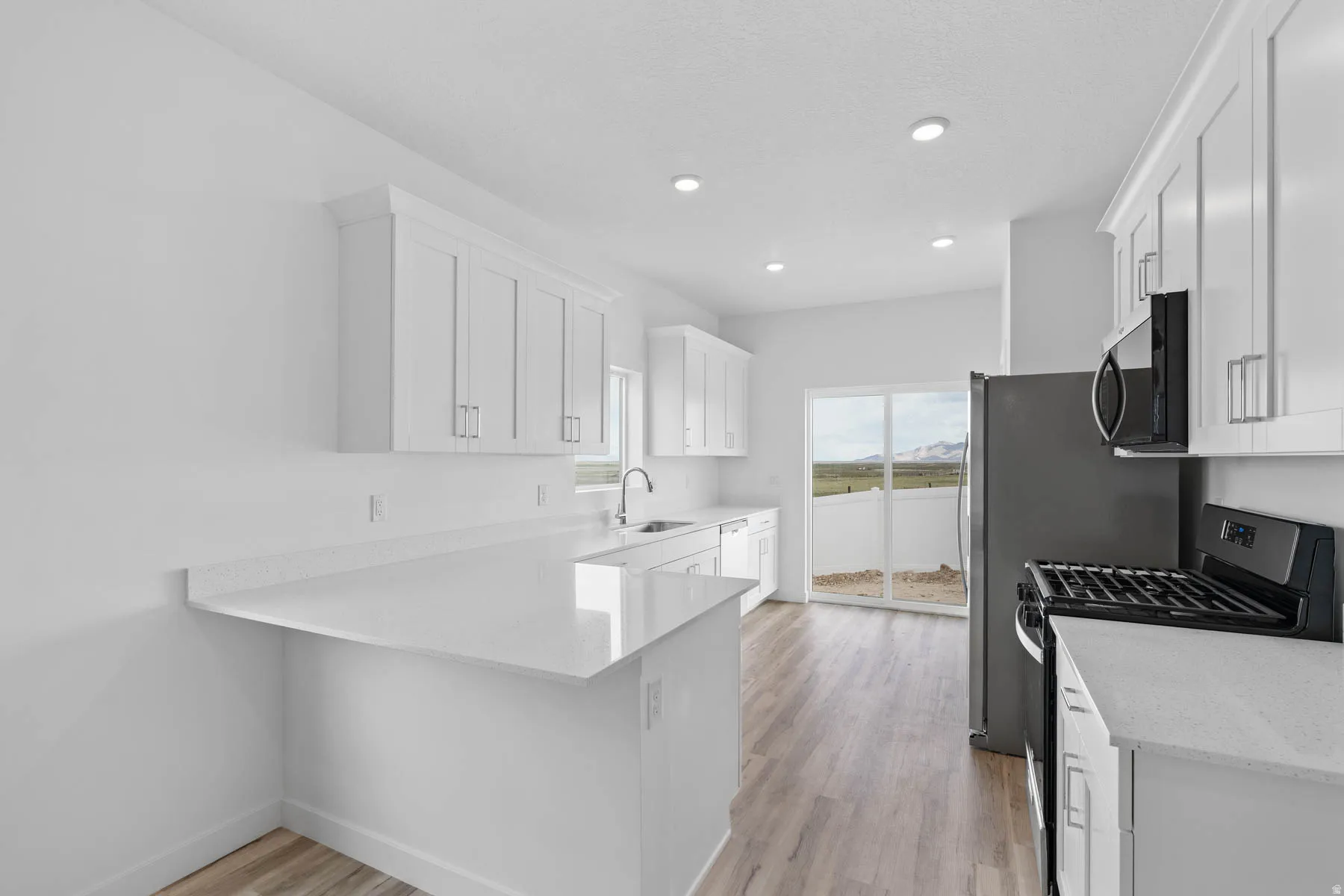 Open-concept kitchen featuring white shaker-style cabinetry, light-toned countertops, wood-finish flooring, a stainless steel refrigerator, and a black gas range