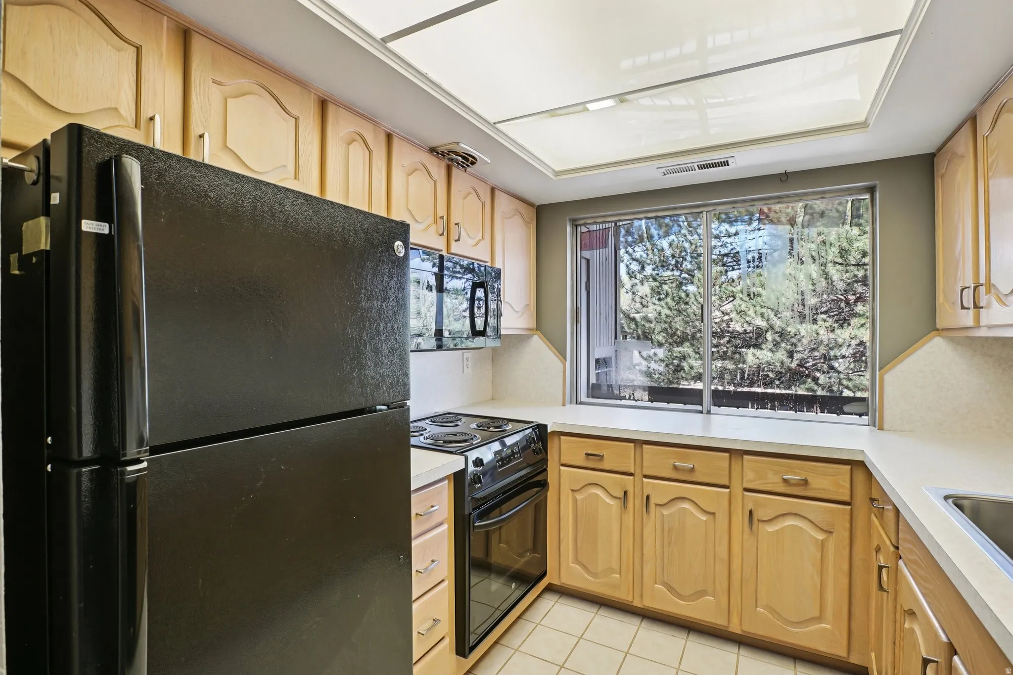 Kitchen with black appliances, light countertops, light wood finish cabinetry, and light tile patterned flooring