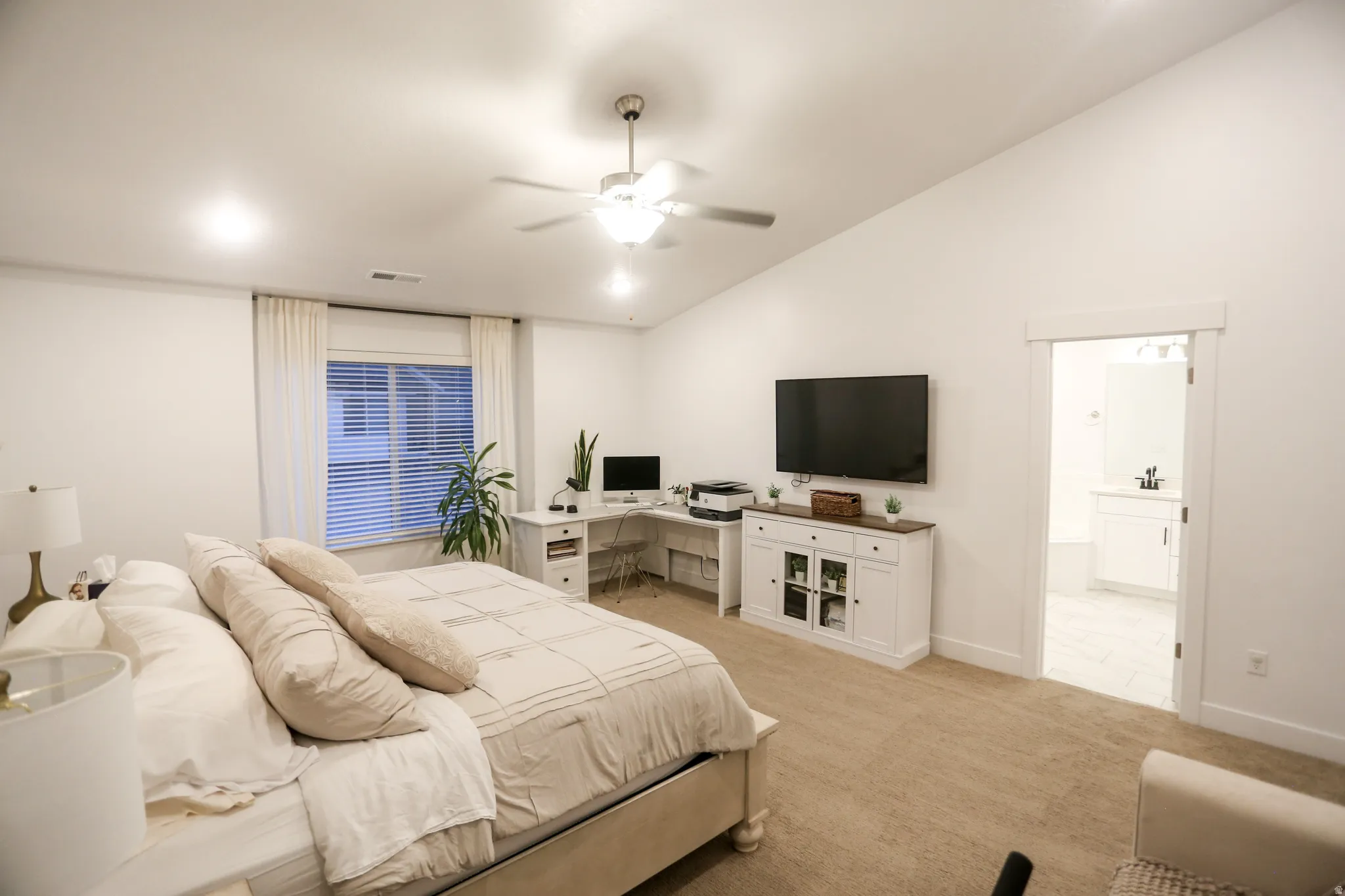 Bedroom with light carpet, vaulted ceiling, ceiling fan, an office area, and connected bathroom