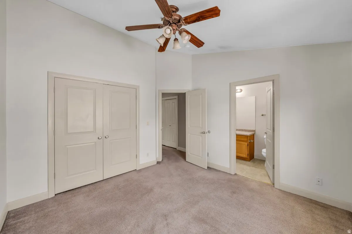 Carpeted room featuring a ceiling fan with wood-finish blades, a vaulted ceiling, and an open doorway revealing a bathroom with a wood-finish vanity