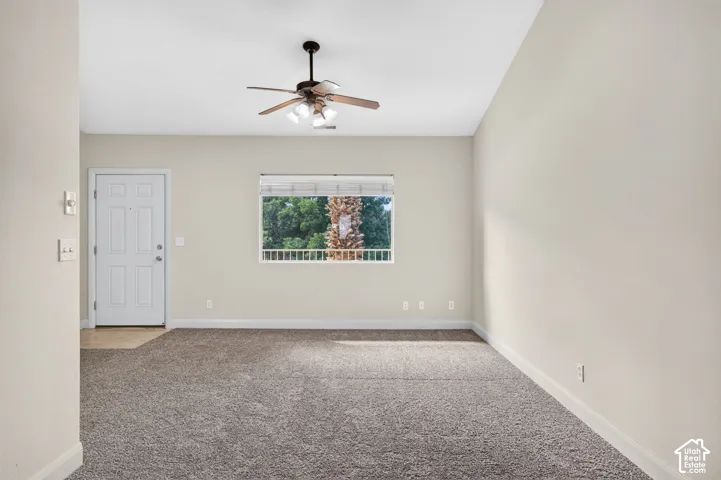 Living space featuring a ceiling fan with integrated lighting, carpeted flooring, a four-panel entry door, and a window with white blinds and exterior landscaping