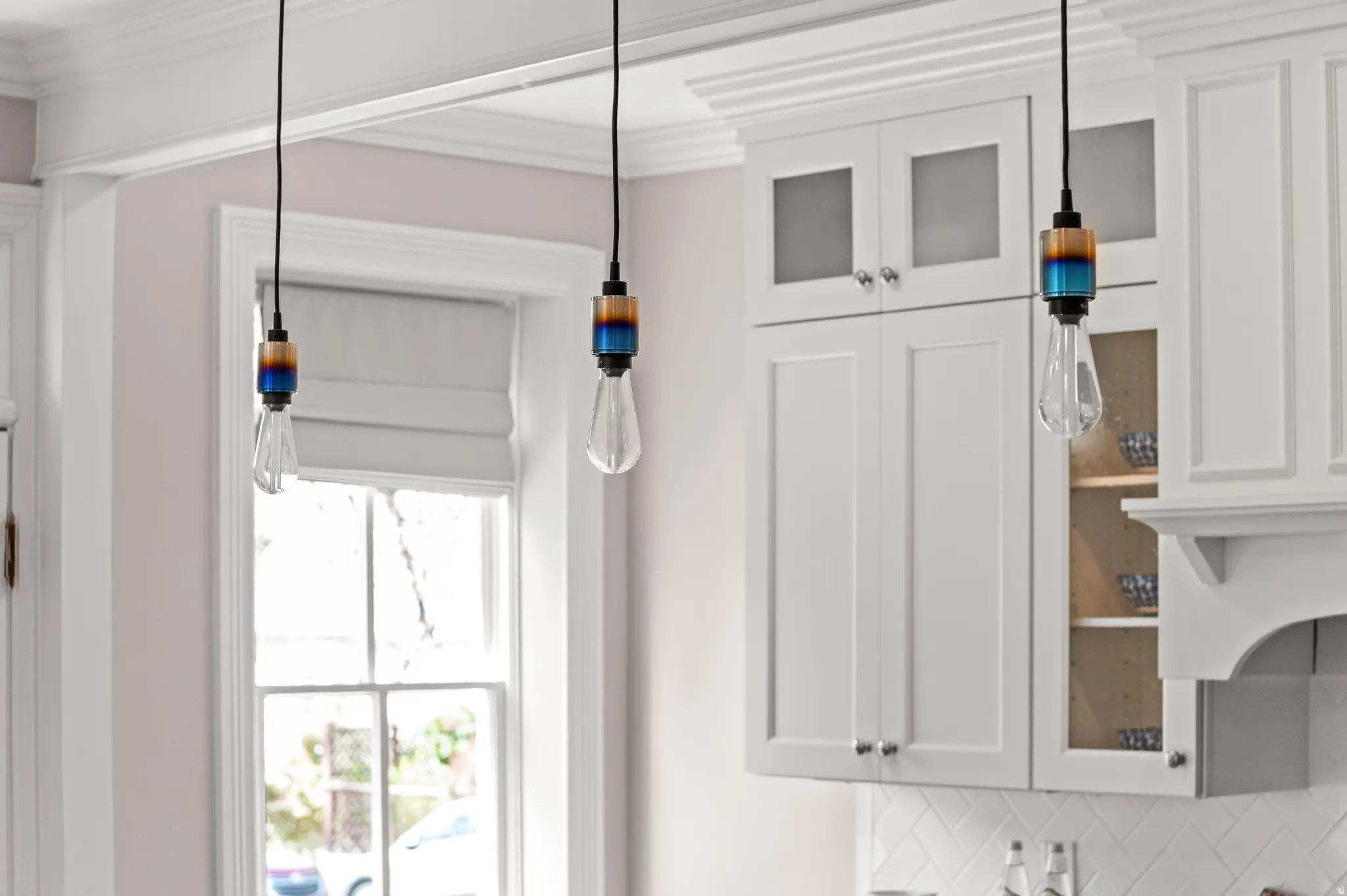 Kitchen view of ornamental molding, white cabinetry, hanging light fixtures, backsplash, and glass fronted cabinets