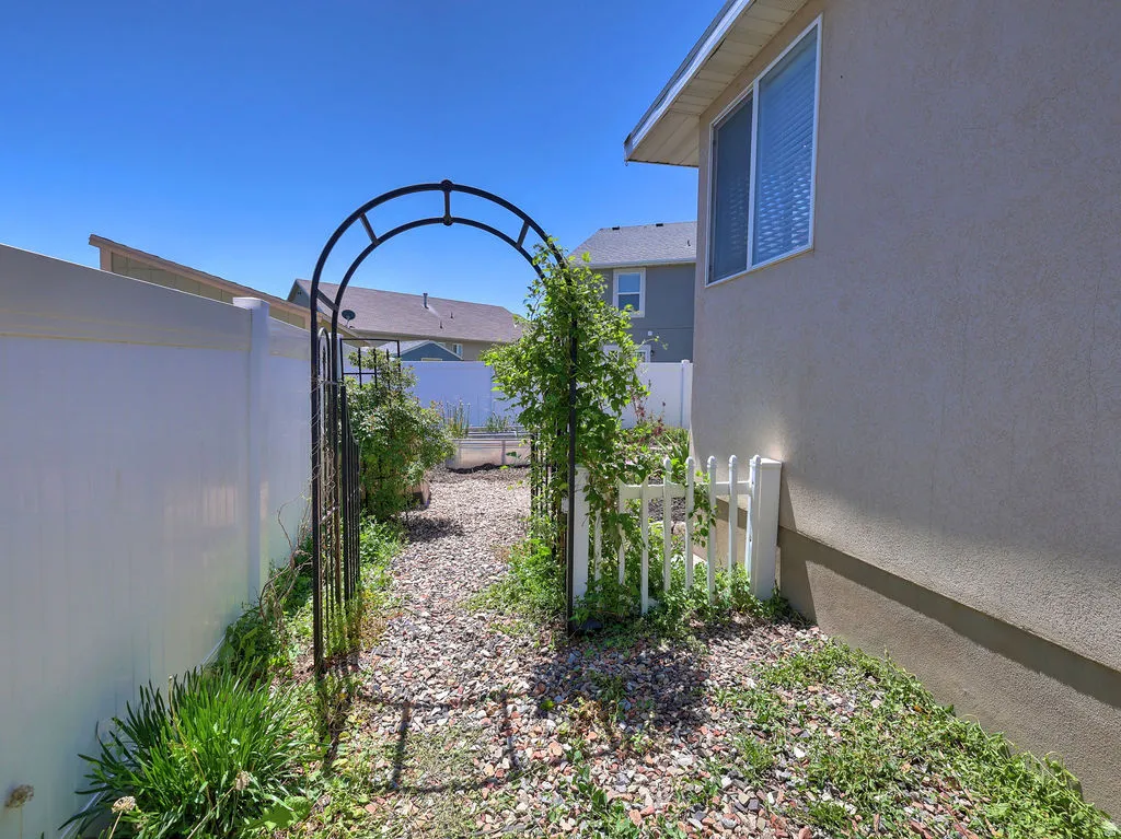 Arched metal garden gate opening to a gravel pathway, framed by a stucco exterior and vinyl fencing