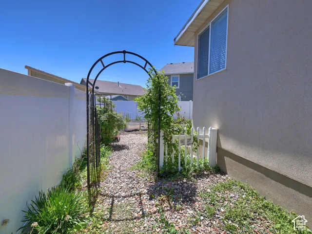 Arched metal garden gate opening to a gravel pathway, framed by a stucco exterior and vinyl fencing