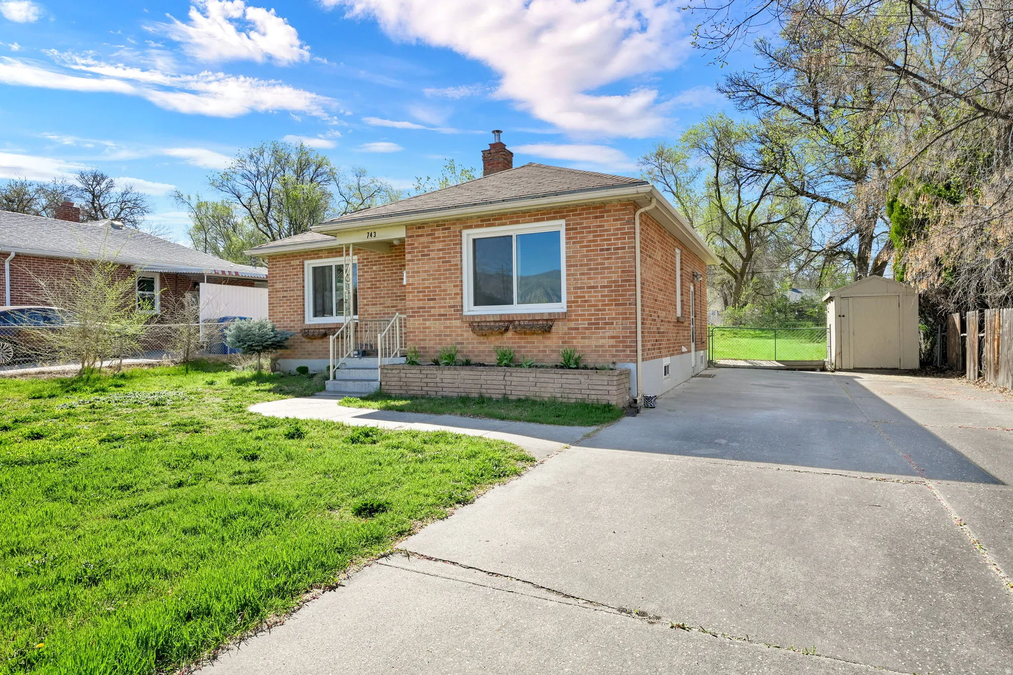 View of front facade with brick siding, driveway, a chimney, a shed, and roof with shingles