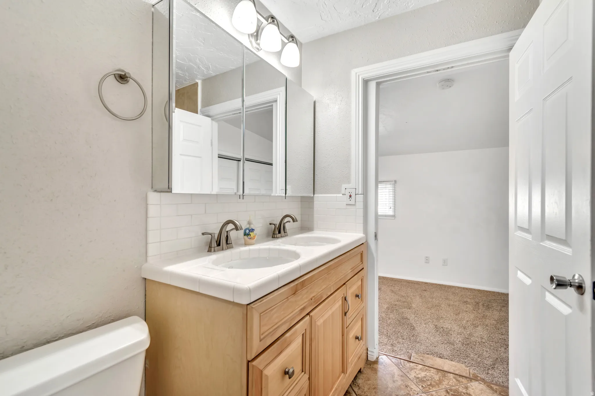 Full bathroom featuring a textured wall, double vanity, decorative backsplash, and light carpet