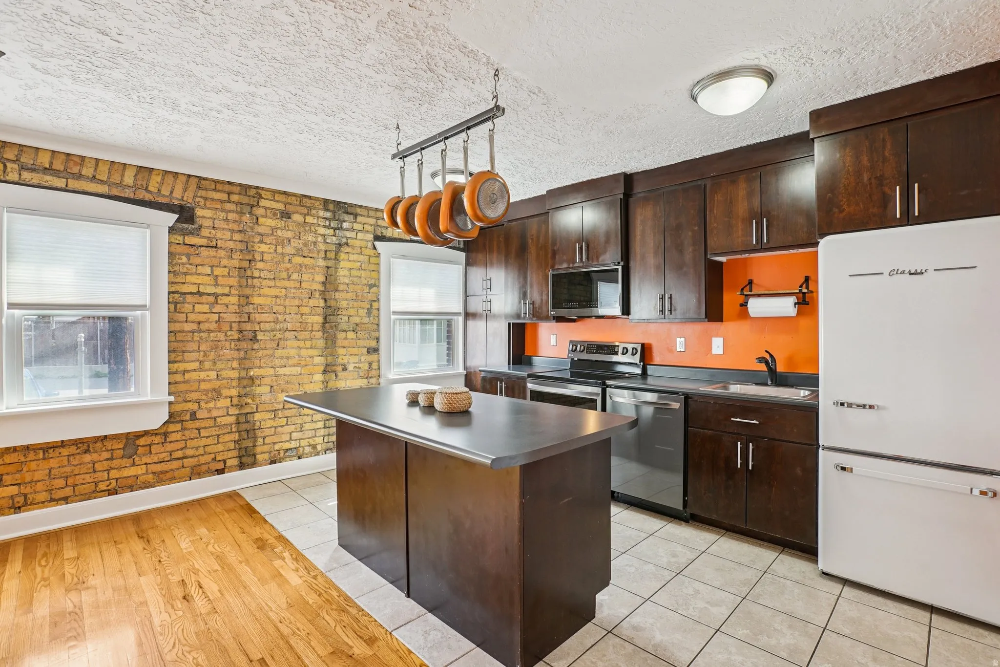 Kitchen with dark wood finish cabinetry, brick wall, stainless steel appliances, light tile patterned flooring, and rail lighting