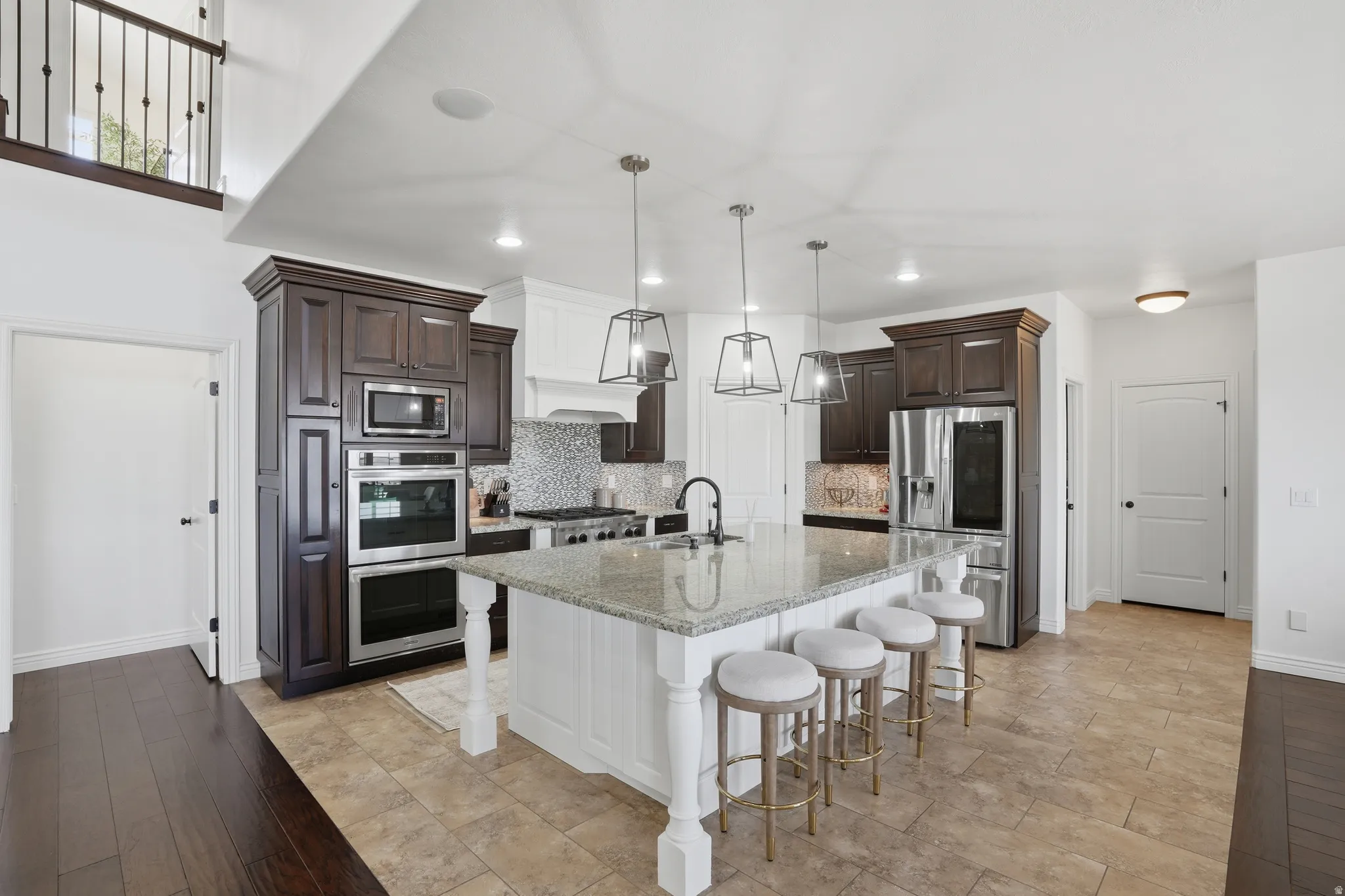 Kitchen with dark wood finish cabinets, light stone countertops, stainless steel appliances, hanging light fixtures, and a center island with sink