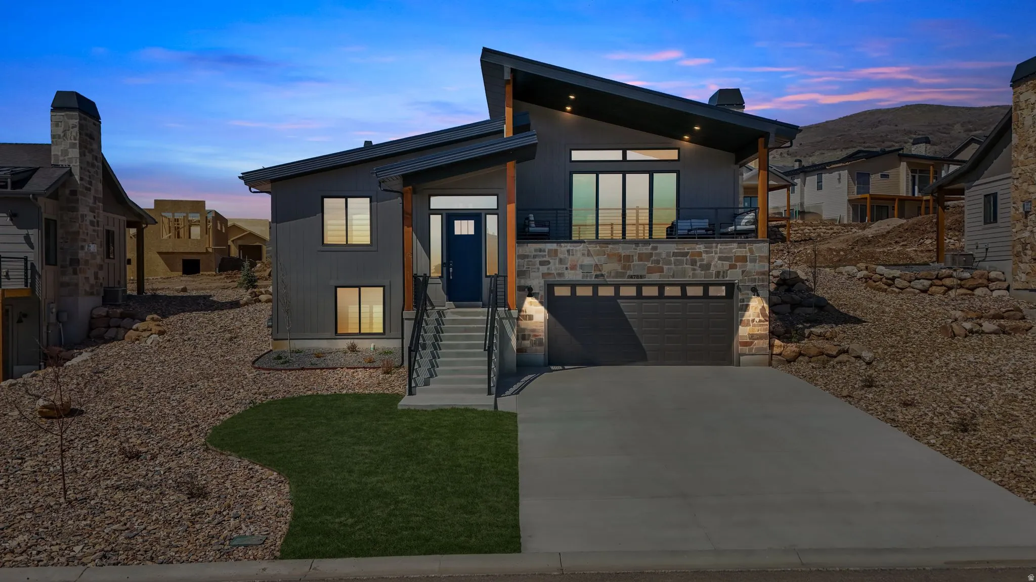Modern architectural exterior featuring a dark gray facade, stone accents, a deep blue entry door, an upper-level balcony, and a two-car garage