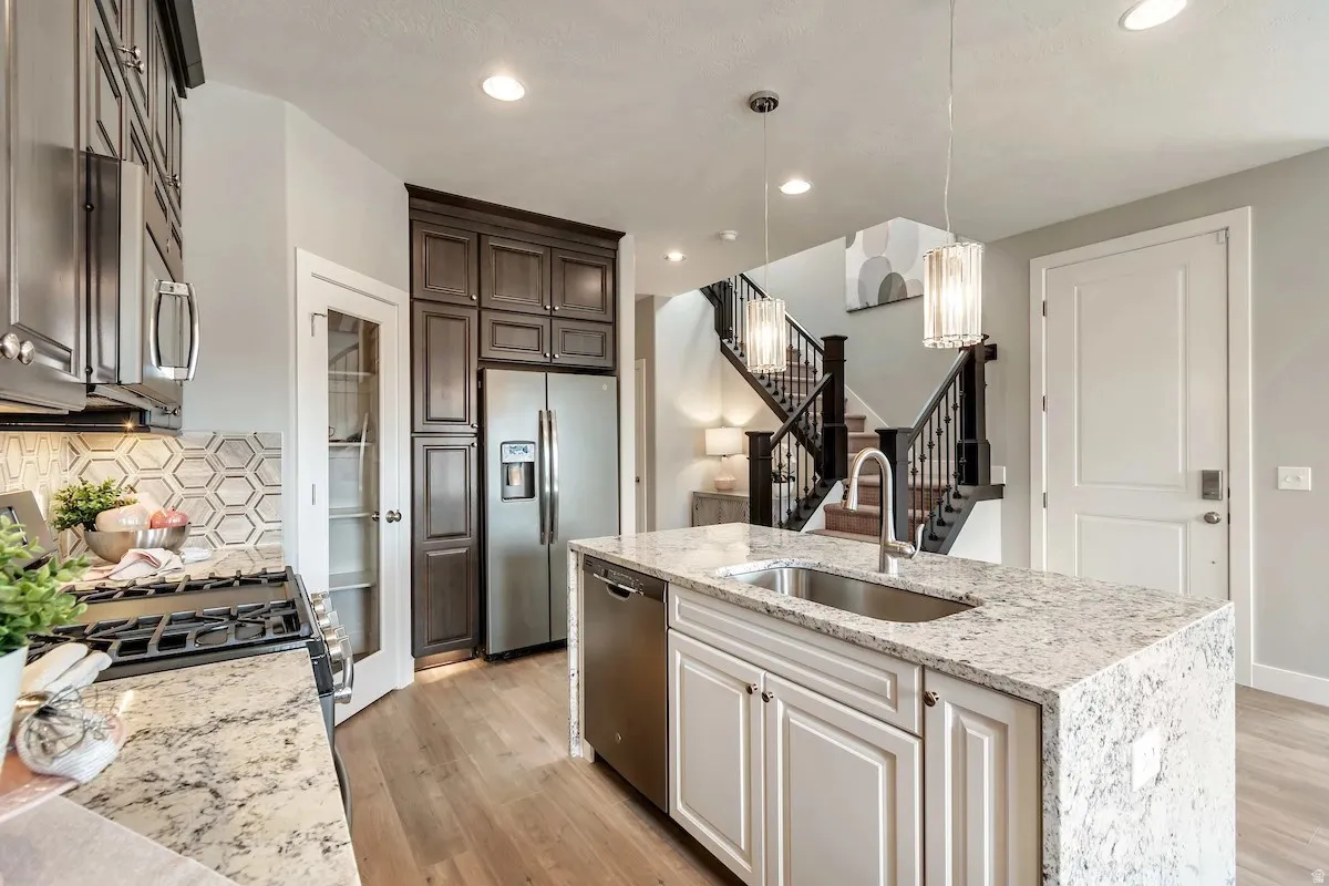 Kitchen with light stone countertops, stainless steel appliances, light wood-type flooring, an island with sink, and decorative light fixtures