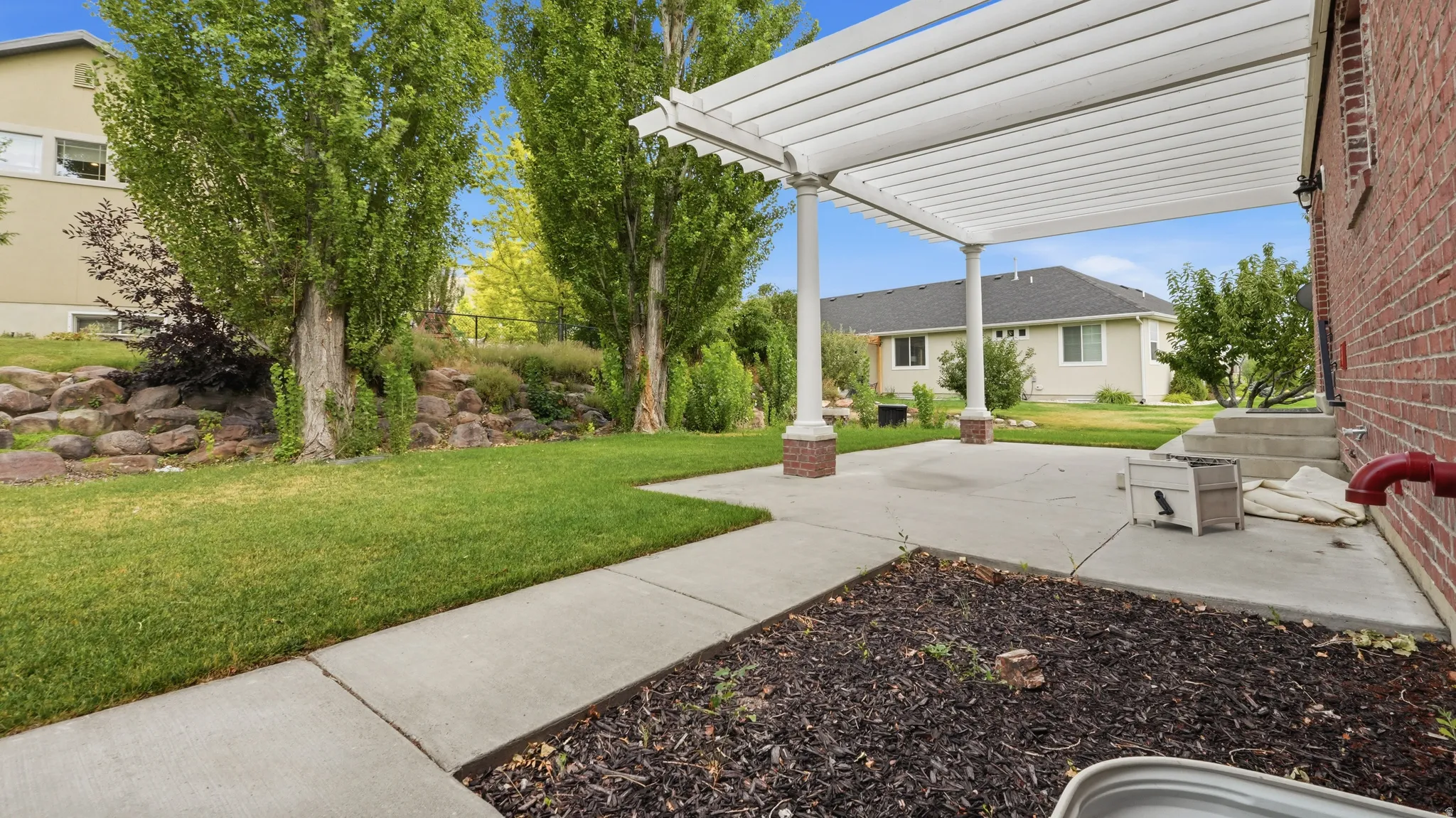 Expansive concrete patio with a white pergola, red brick exterior, and a manicured lawn