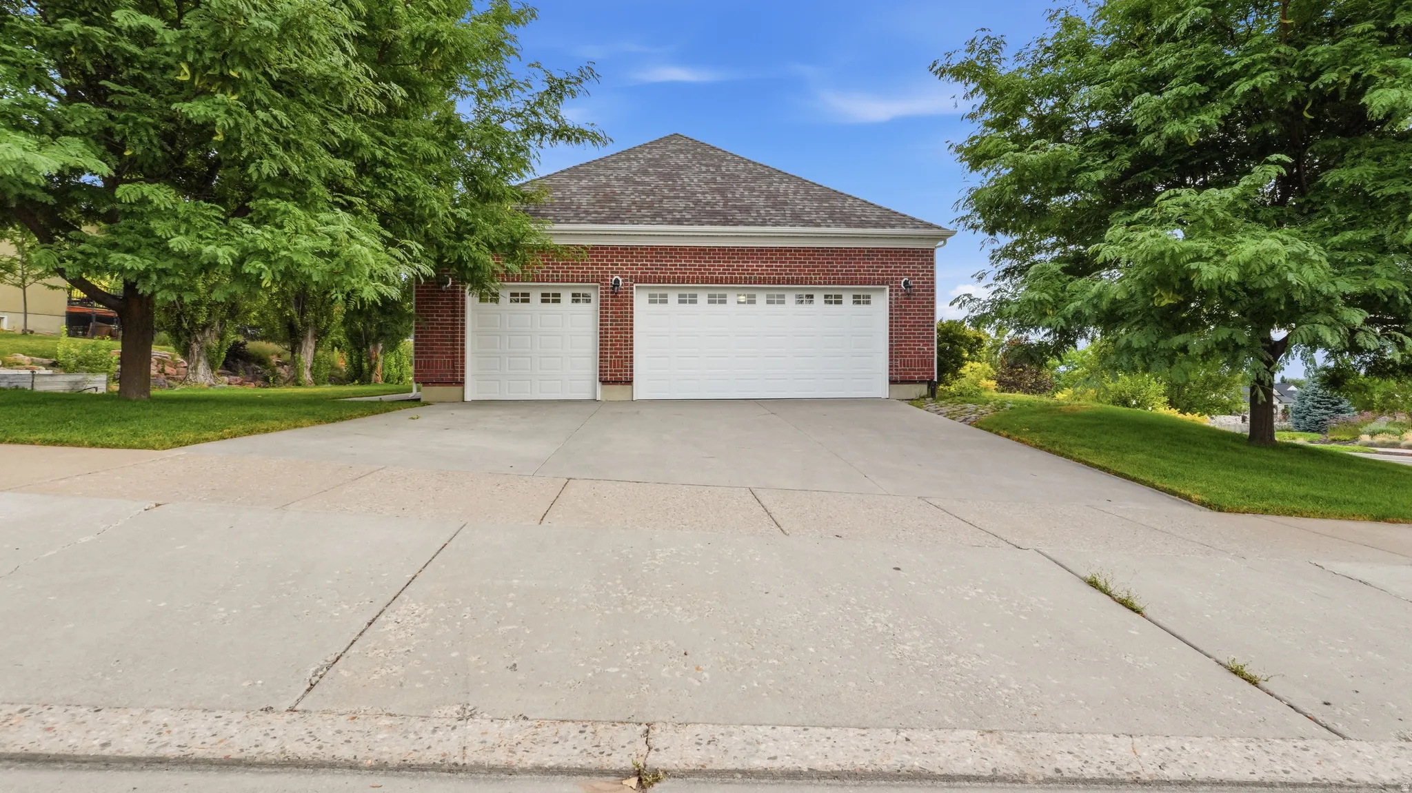 Brick exterior with a peaked roofline, two white garage doors, a concrete driveway, and established trees