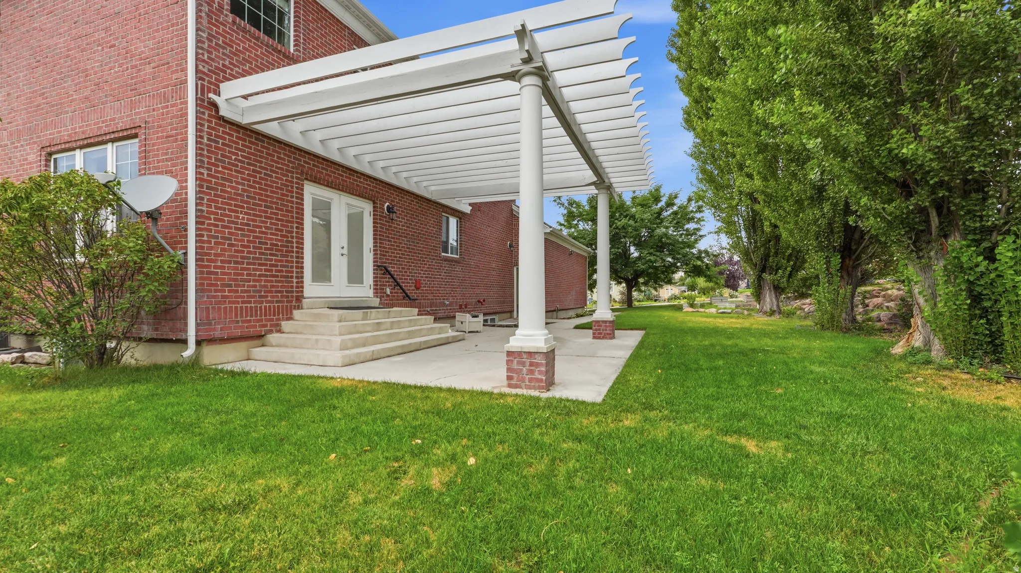 Brick Colonial home featuring a spacious rear patio with attached pergola structure, manicured lawn, and mature landscaping including established trees and hedging