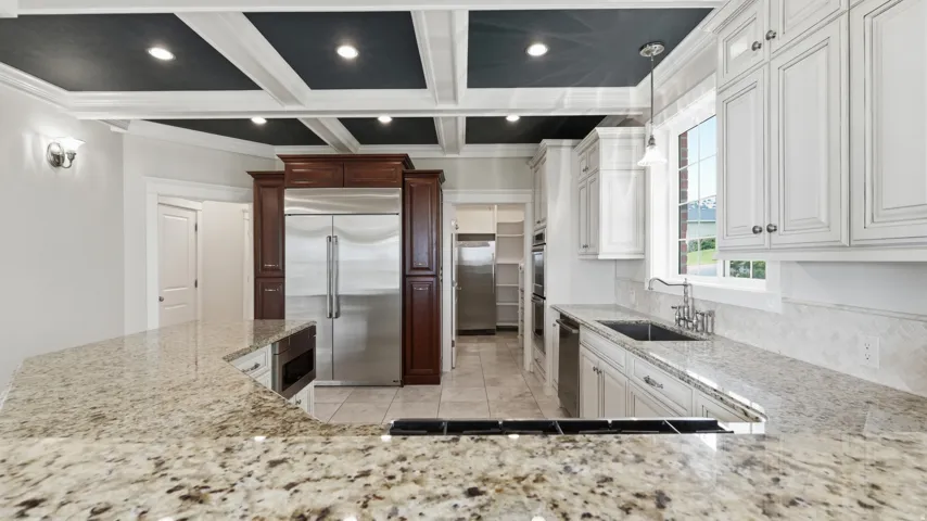 Spacious kitchen featuring white cabinetry with hardware accents, granite countertops, stainless steel appliances, a large center island with seating, and coffered ceiling with recessed lighting