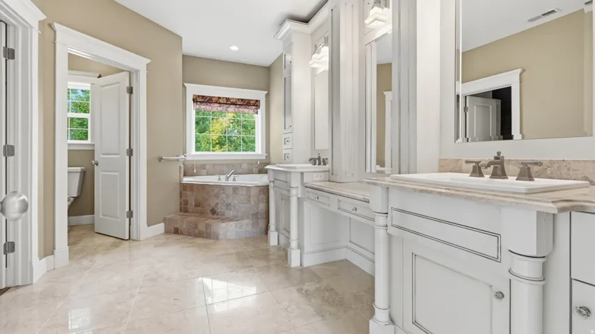 Luxurious primary bathroom suite featuring dual vanities with polished stone countertops, an oversized soaking tub with tile surround, and abundant natural light from multiple windows