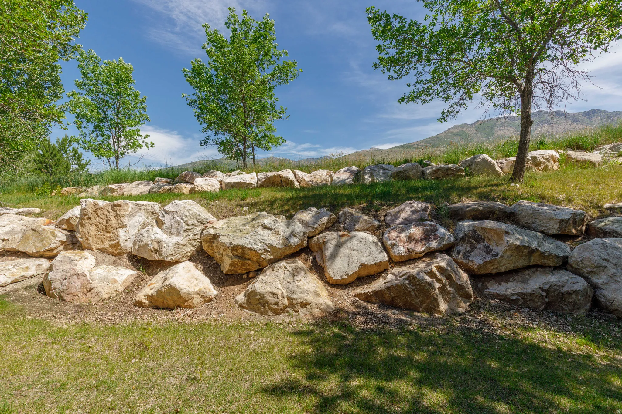 Layered rock retaining wall with natural stone elements, grassy terrain, mature trees, and distant mountain range