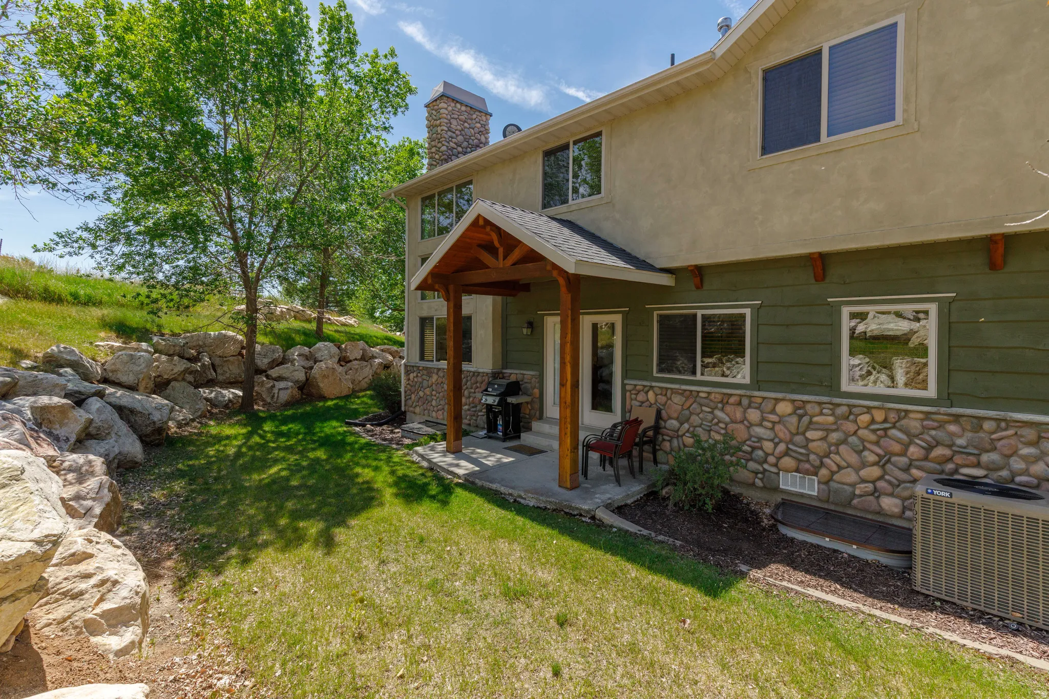 Rear exterior featuring a covered patio with exposed wood beams, a stone-clad foundation, a stucco upper level, and a sloped lawn with large rock landscaping