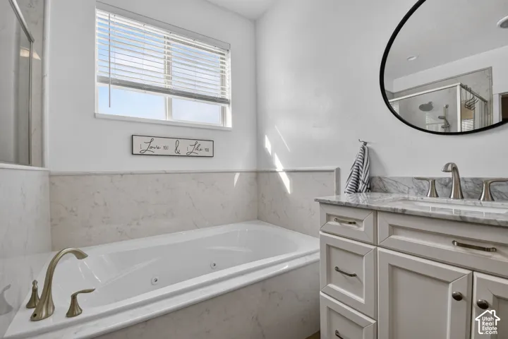 Soaking tub with brass-finish fixtures, a large window with blinds, and marble-finish tile surround
