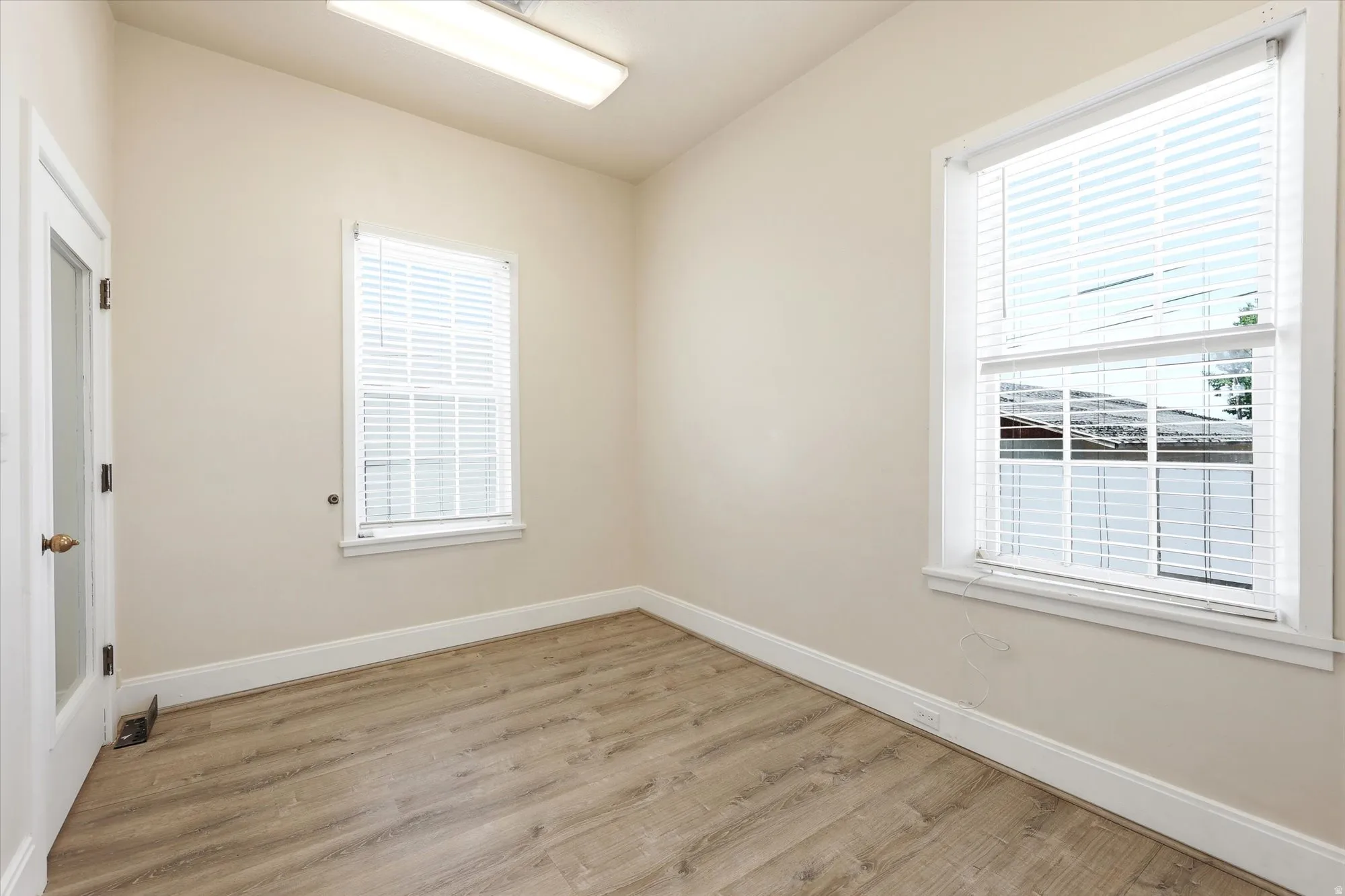 Room featuring two windows with white blinds, wood-finish flooring, light neutral-toned walls, and a glass-paneled door