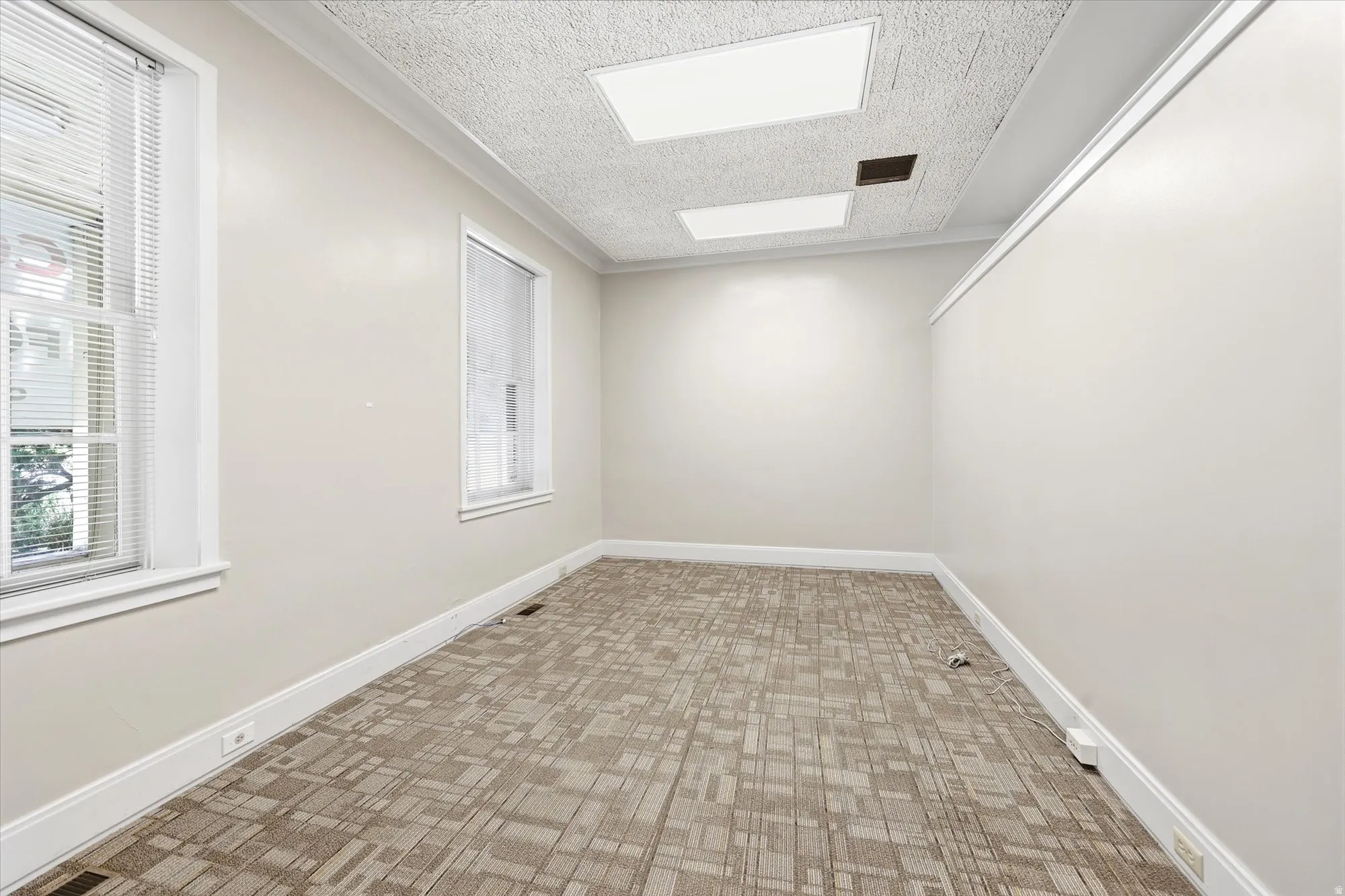 Neutral-toned room featuring two windows with white blinds, textured carpet flooring, white baseboards, and a dropped ceiling with recessed lighting