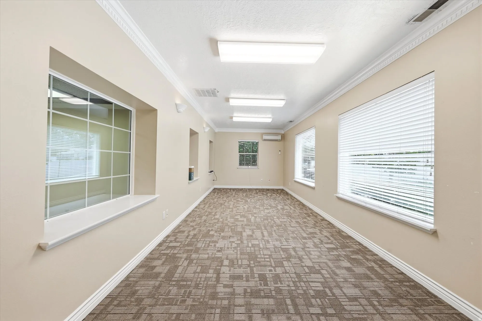 Spacious room featuring patterned carpet flooring, multiple windows with blinds, recessed wall niches, crown molding, and fluorescent ceiling lights