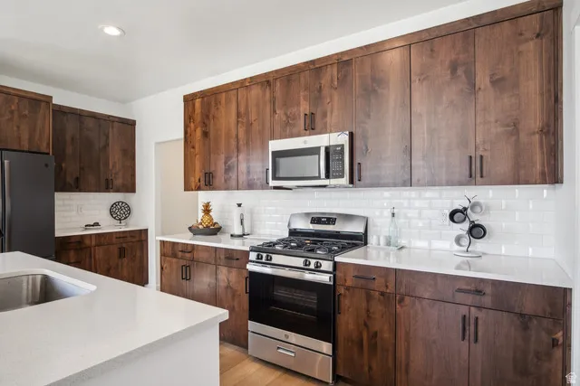 Kitchen with stainless steel appliances, dark wood finish cabinetry, light wood-style flooring, decorative backsplash, and light stone countertops