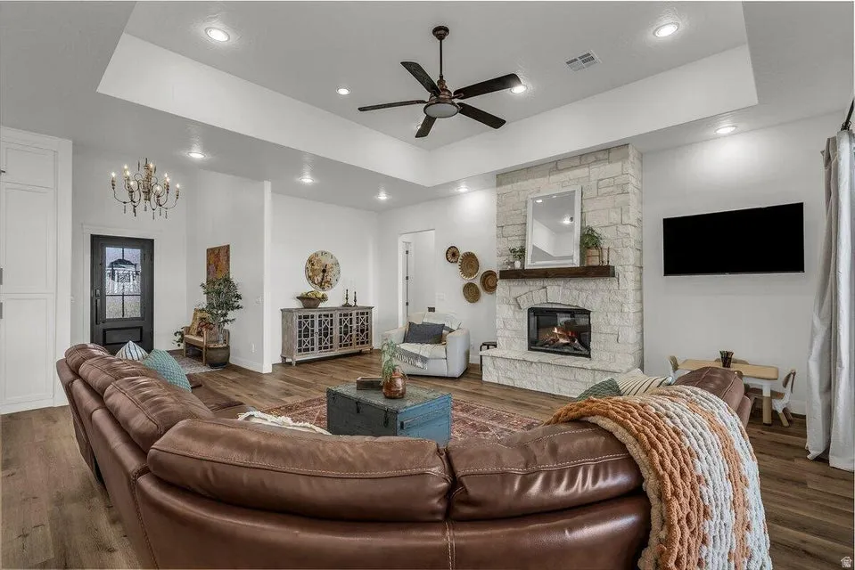 Living area with a raised ceiling, ceiling fan, dark wood-type flooring, a stone fireplace, and a chandelier