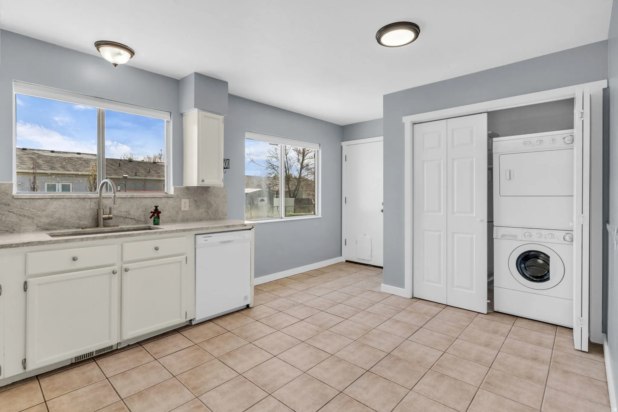 Kitchen with white cabinetry, stacked washing machine and dryer, light stone counters, dishwasher, and decorative backsplash