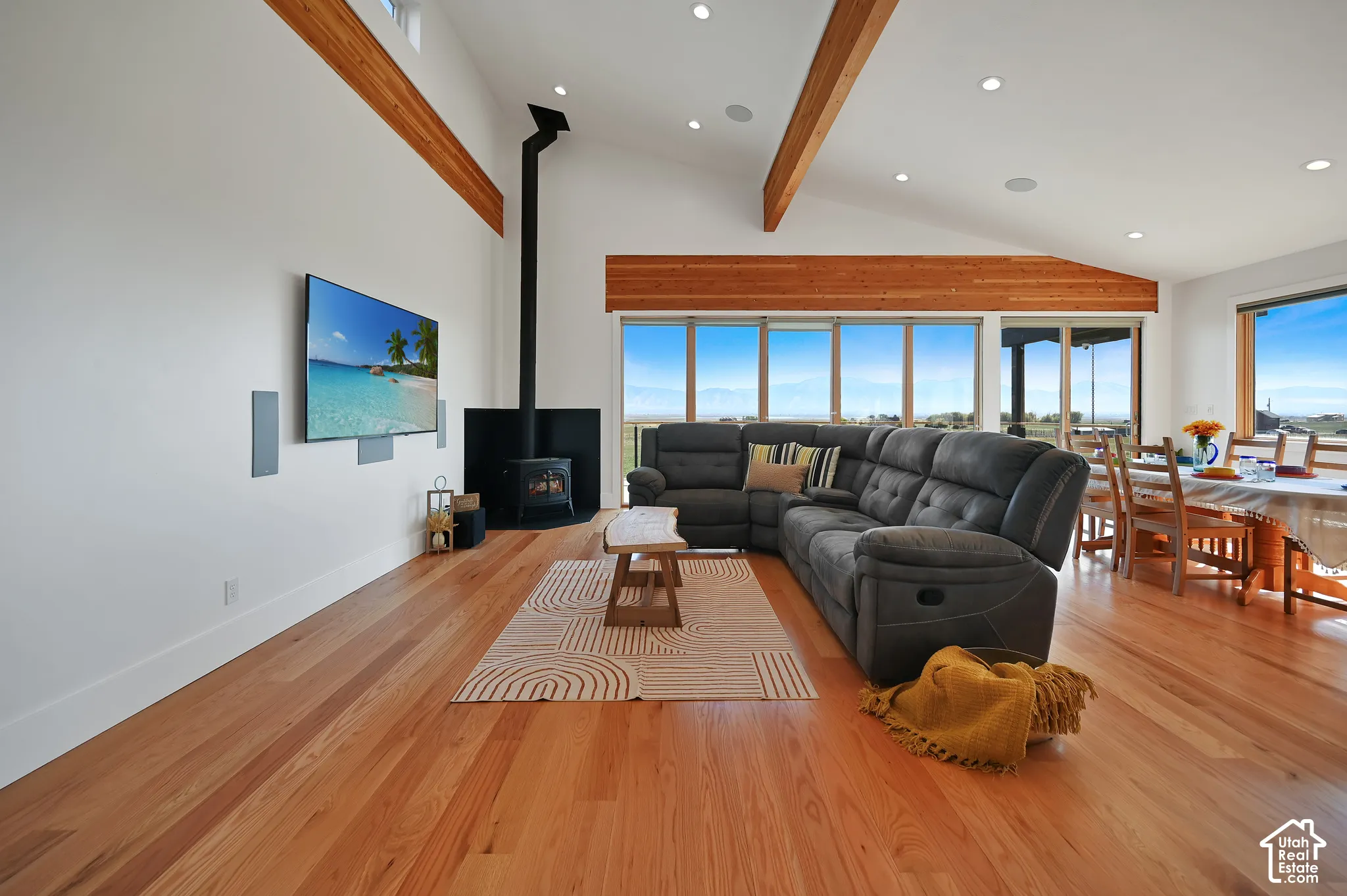 Vaulted ceilings with exposed wood beams, wood-finish flooring, recessed lighting, and a corner wood-burning stove with a black surround