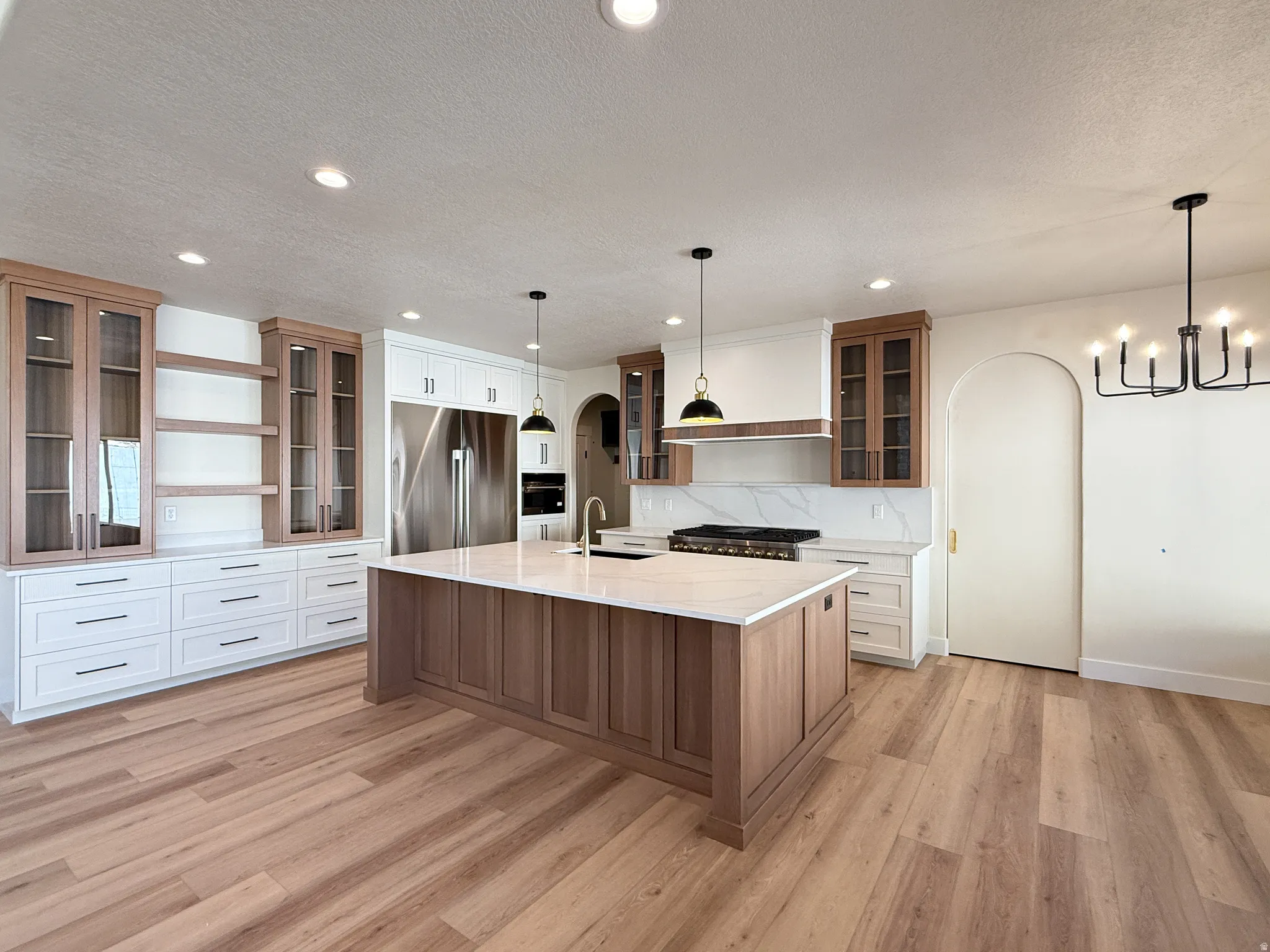 Two tone kitchen with glass fronted cabinets, open shelves, arched walkways, stainless steel appliances, and a kitchen island with sink