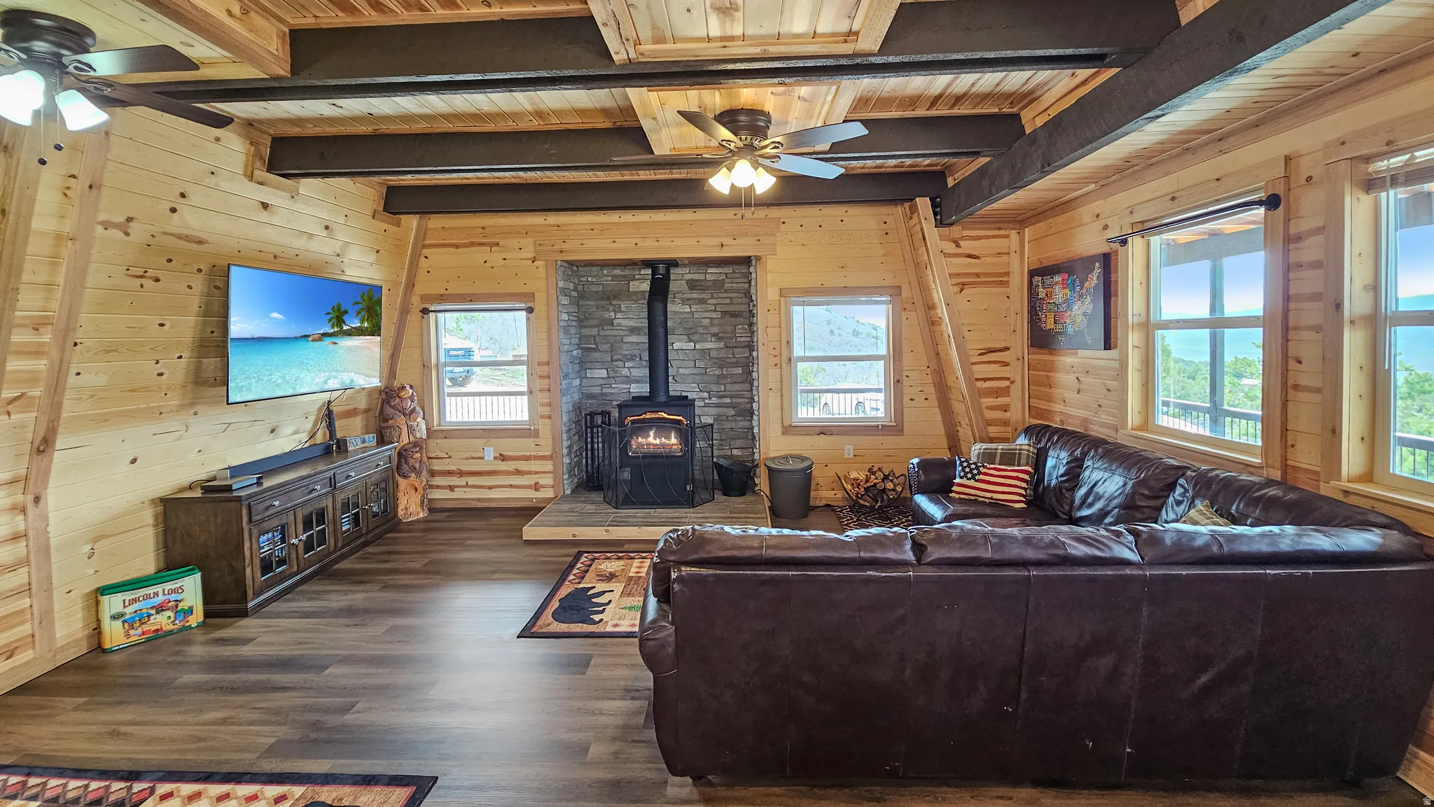 Living area featuring ceiling fan, a wood stove, wood walls, dark wood-type flooring, and a wood ceiling with exposed beams