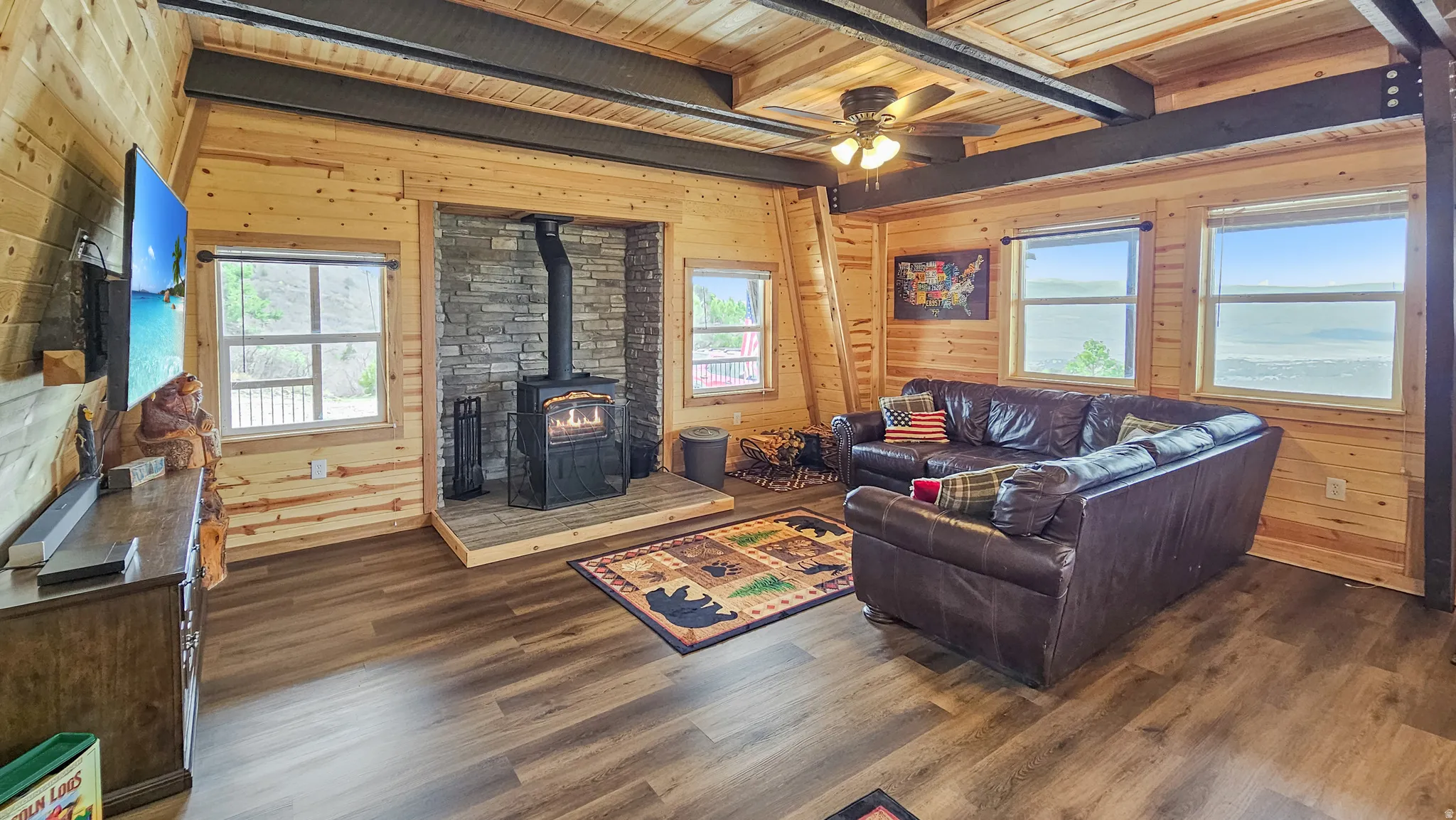 Living room with wood walls, a wood stove, a wooden ceiling with exposed beams, dark wood finished floors, and ceiling fan