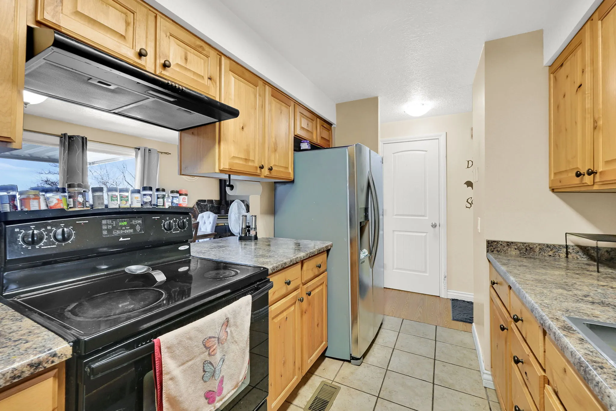Kitchen with electric range, range hood, stainless steel fridge with ice dispenser, light tile patterned floors, and light wood finish cabinets