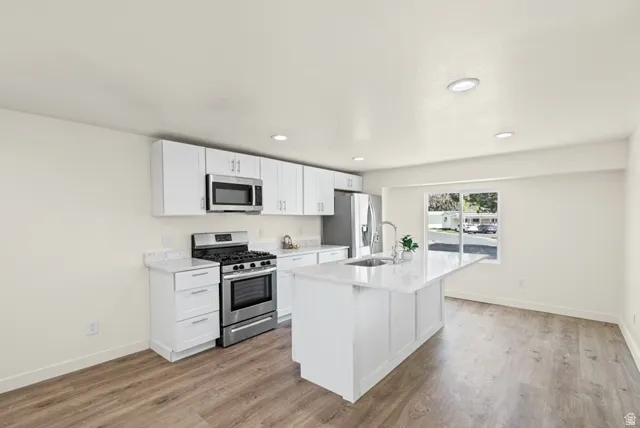Kitchen with stainless steel appliances, white cabinetry, light wood-type flooring, a center island with sink, and light stone countertops