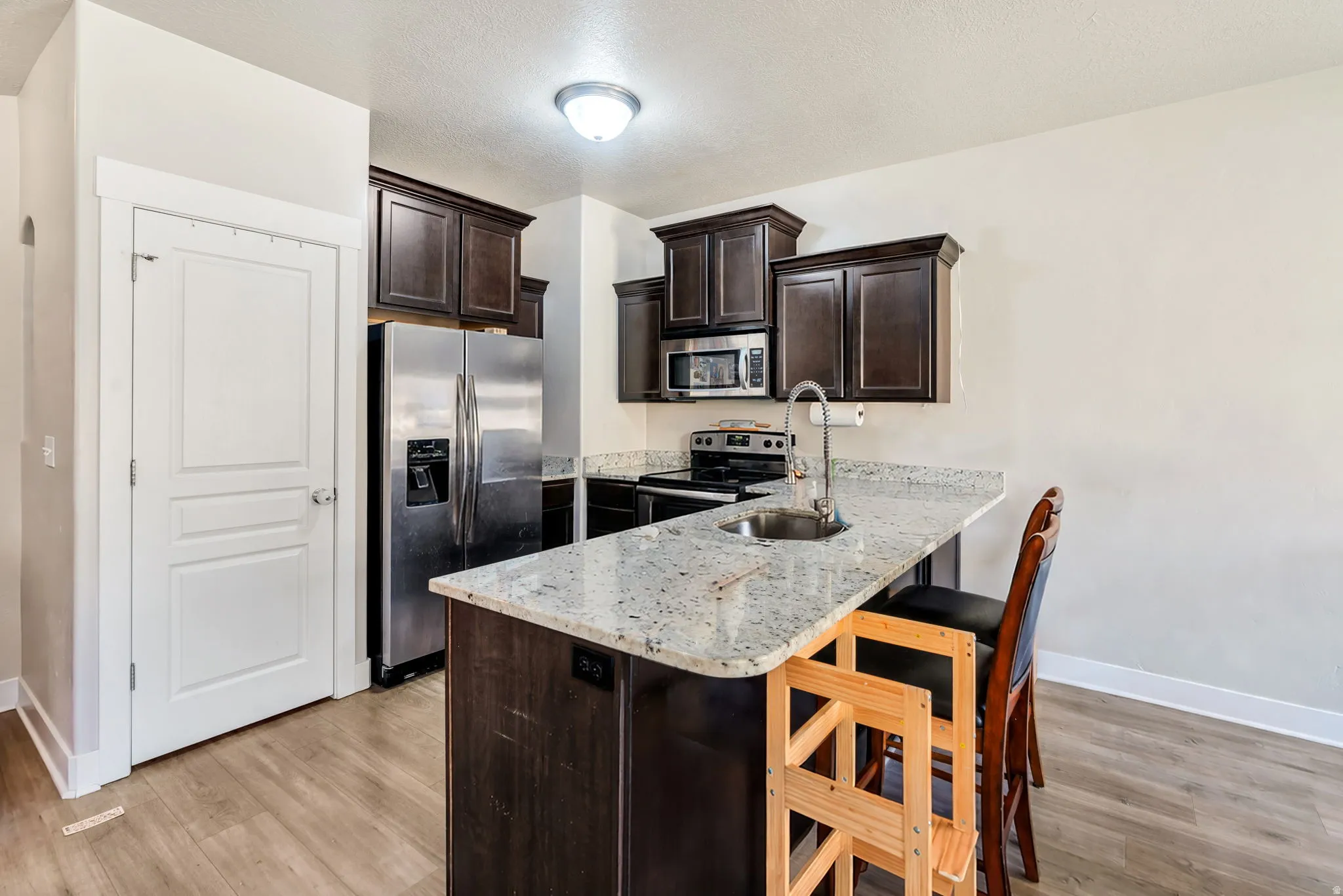 Kitchen featuring dark wood finish cabinetry, stainless steel appliances, a peninsula, light stone countertops, and a kitchen bar
